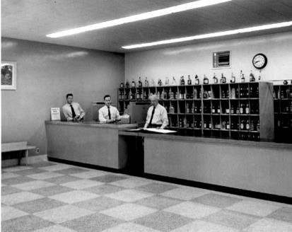 Une photo en noir et blanc d’un ancien style de magasin. Trois commis de sexe masculin sont derrière le comptoir et on peut apercevoir derrière eux des tablettes remplies de bouteilles.