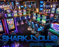 Une pièce sombre plus éclairée grâce à des appareils de loterie vidéo. On peut lire le texte : « Shark Club gaming centre ».