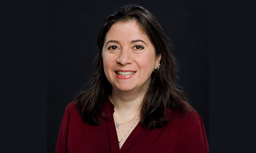 A woman in front of a dark background is wearing a burgundy blouse, two gold necklaces, and hoop earrings. She is smiling at the camera.