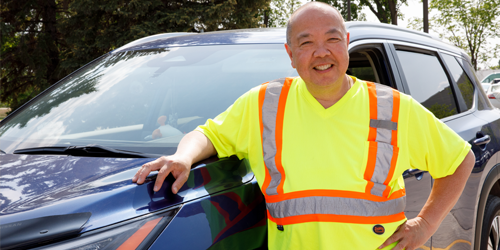 A smiling man wearing a bright yellow high-visibility safety shirt with orange and silver reflective stripes, standing beside a blue SUV in an outdoor parking lot surrounded by trees. The man is leaning on the vehicle with one hand and has the other hand on his hip.